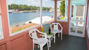 A covered outdoor area with a view of a lake, featuring white plastic chairs, a table with a bottle and glass, and a sign with the word "BEACH". - The Naswa Resort (Laconia)