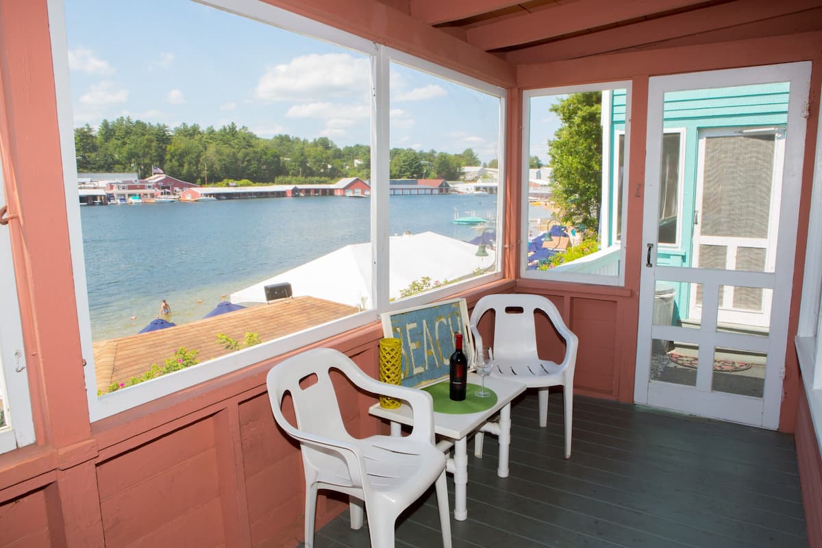A covered outdoor area with a view of a lake, featuring white plastic chairs, a table with a bottle and glass, and a sign with the word "BEACH".