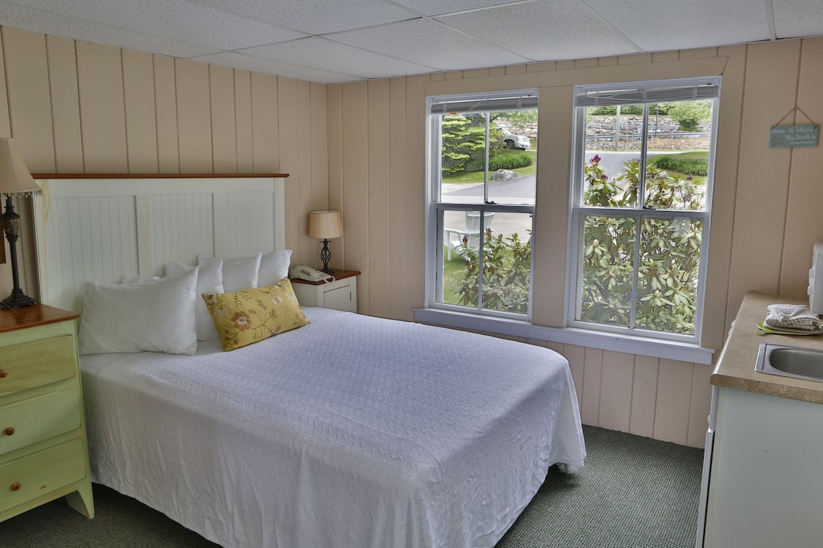 A neatly arranged bedroom with a large bed, a nightstand, a window with a view of the outside, and a small kitchenette area.