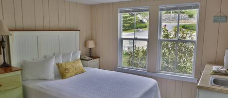 Un dormitorio ordenado con una cama grande, una mesita de noche, una ventana con vistas al exterior y una pequeña zona de kitchenette.