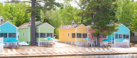 A row of colorful beach cottages with outdoor seating and umbrellas, situated by a body of water.
