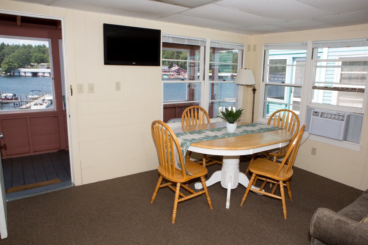 A dining area with a table and chairs, a television mounted on the wall, and windows with a view of the water and boats.