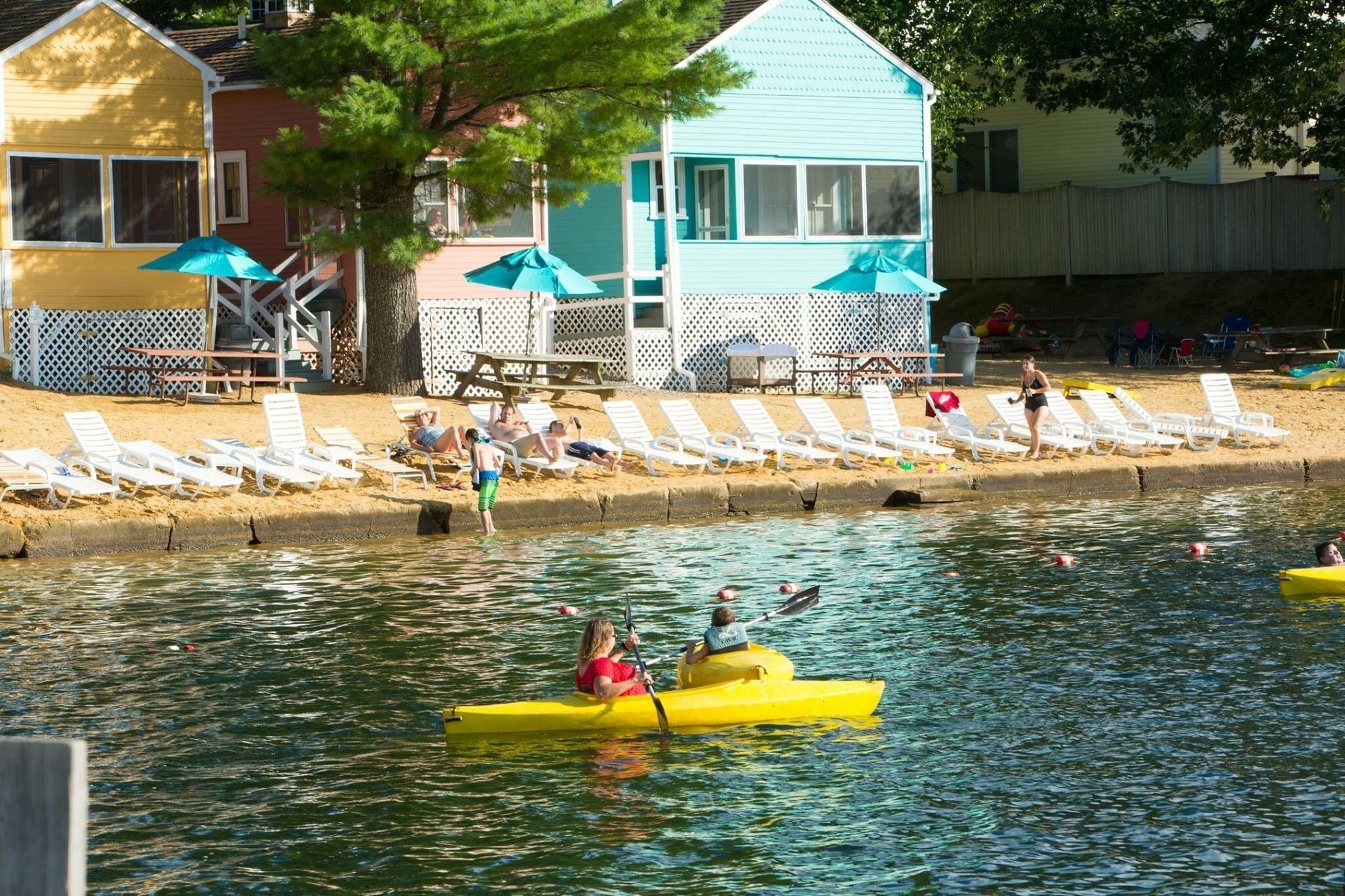 A lakeside setting with a yellow and blue building, white deck chairs, and a kayak on the water.