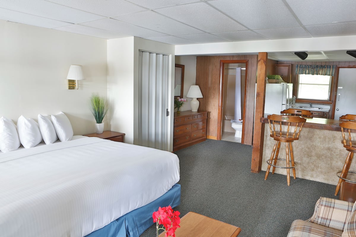 A hotel room with a large bed, a desk, a chair, a small table with a vase, and a kitchenette area.