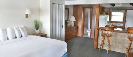 A hotel room with a large bed, a desk, a chair, a small table with a vase, and a kitchenette area.