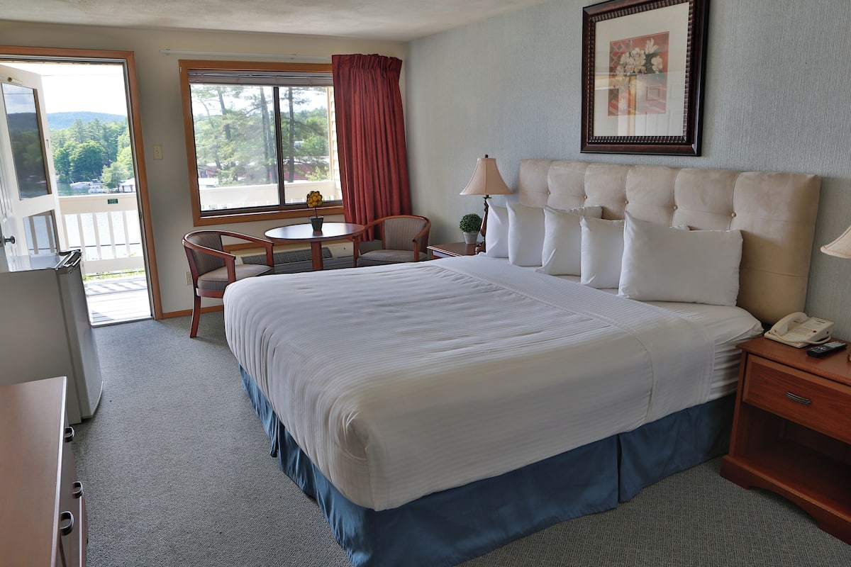 A hotel room with a large bed, a desk, a chair, a small table, a lamp, and a window with a view of trees and a building.