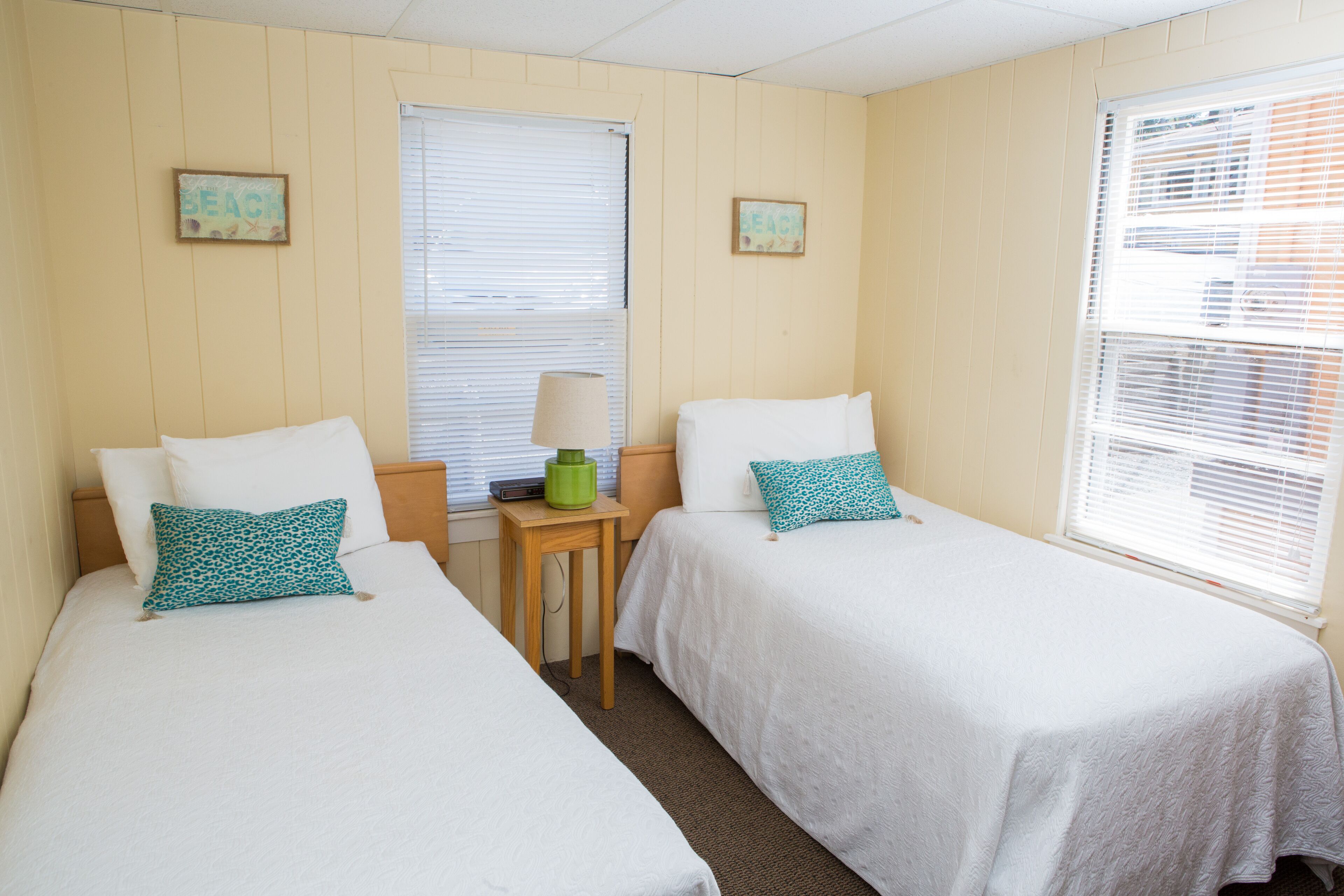 A room with two single beds, each with white bedding and a green patterned pillow. There is a small wooden nightstand with a lamp between the beds. The walls are beige, and there are two framed pictures on the wall above the beds.