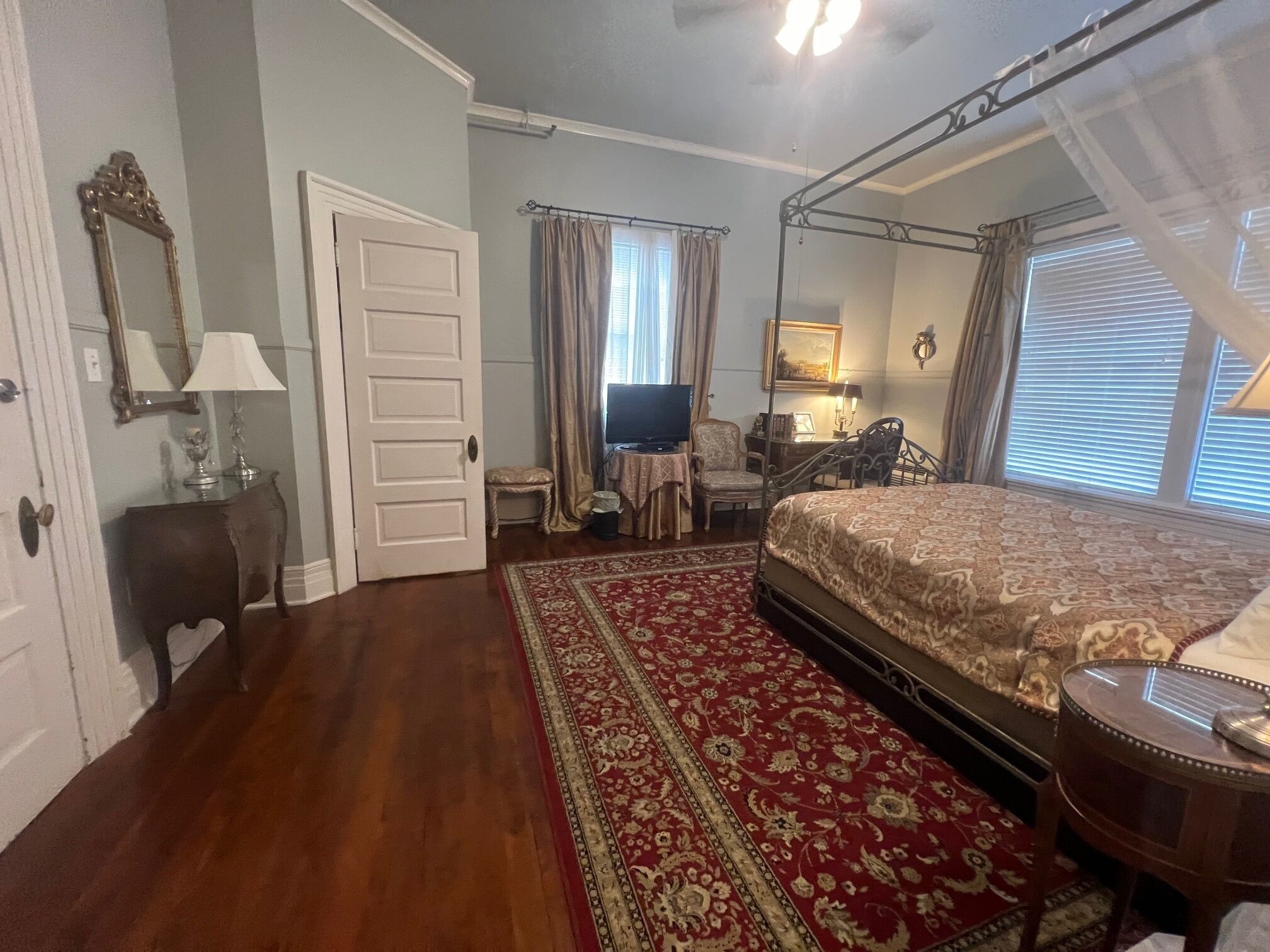 A bedroom with a four-poster bed, a television, a desk with a chair, and a large window with curtains.