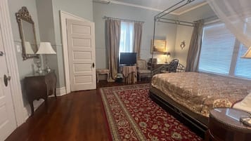 A bedroom with a four-poster bed, a television, a desk with a chair, and a large window with curtains.