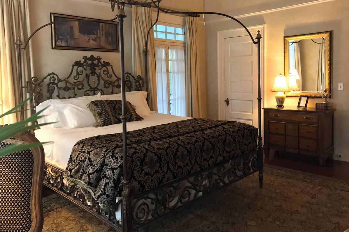 Una cama con dosel, con un edredón oscuro de estampado, un cabecero de madera y una mesita de noche con lámpara.