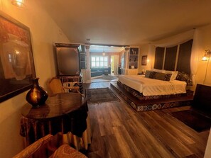 A bedroom with a large bed, a desk, and a chair. There is a window with curtains and a view of the outside.