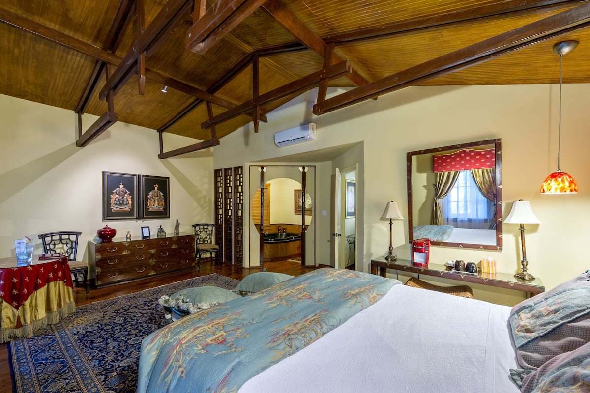 Un dormitorio con una cama grande, techo de madera, cómoda, espejo y una mesa roja con un vaso.