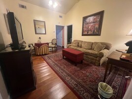 Una sala de estar con un sofá, un reposapiés rojo, un televisor sobre un mueble, una mesa de comedor con sillas y una alfombra estampada.