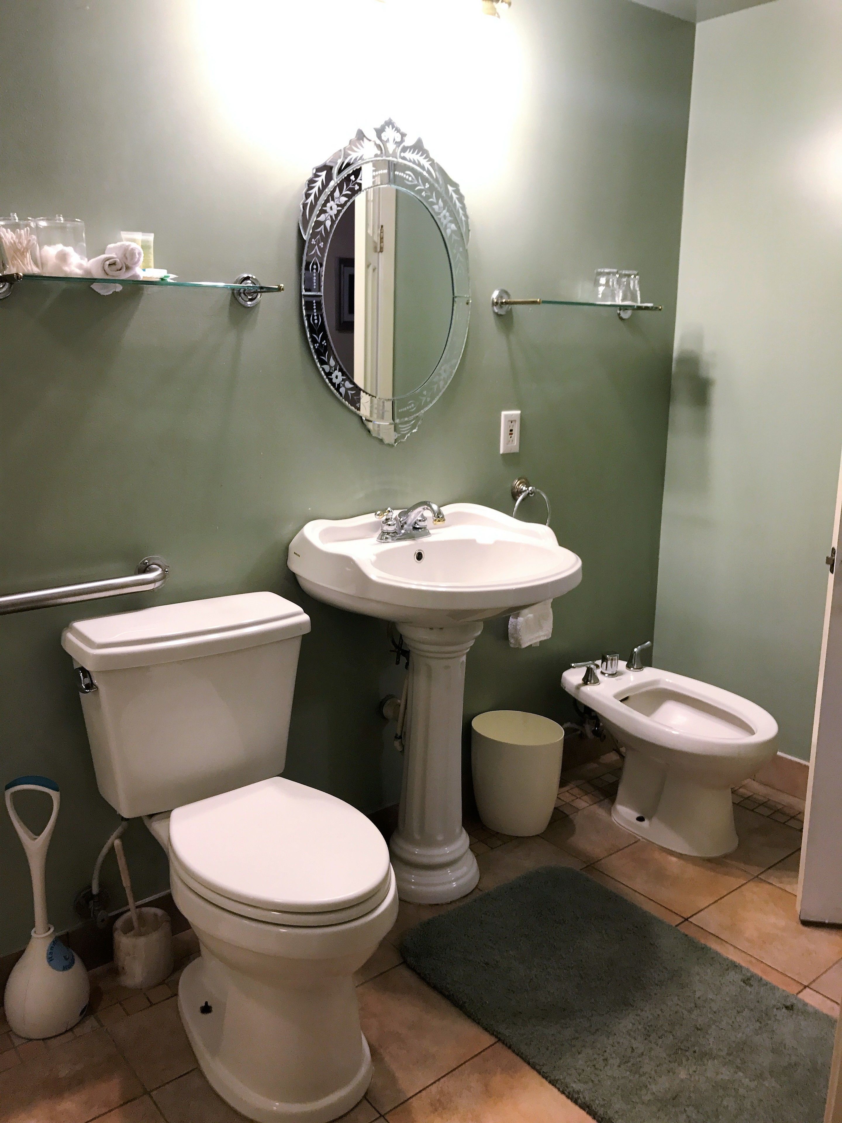 Un baño con inodoro, bidé, lavamanos pedestal y espejo con marco decorativo.