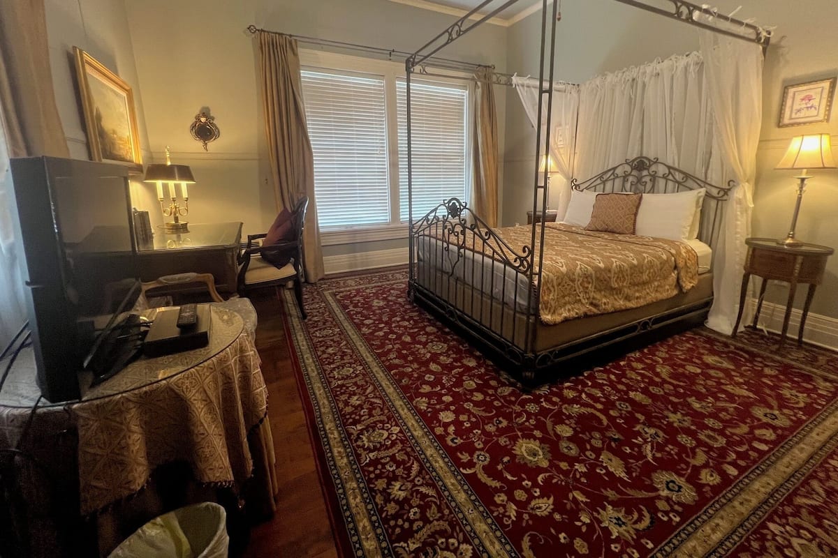 Una cama con dosel y cuatro columnas con una colcha de diseño rojo y dorado, un televisor, una mesita con lámpara y una silla en una habitación con alfombra roja y un gran ventanal con cortinas.
