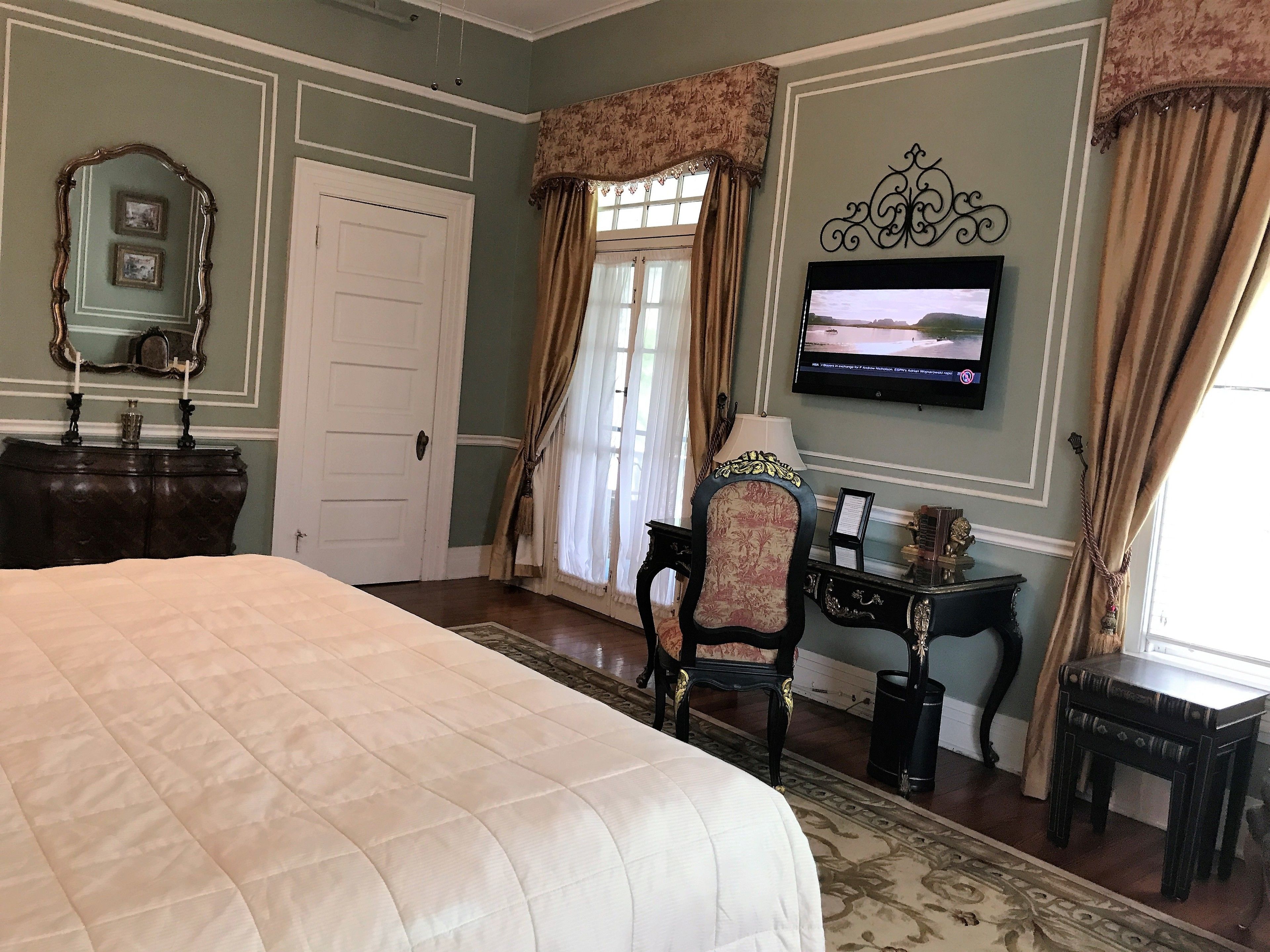 A bedroom with a large bed, a television on the wall, a chair, a desk, and a door.