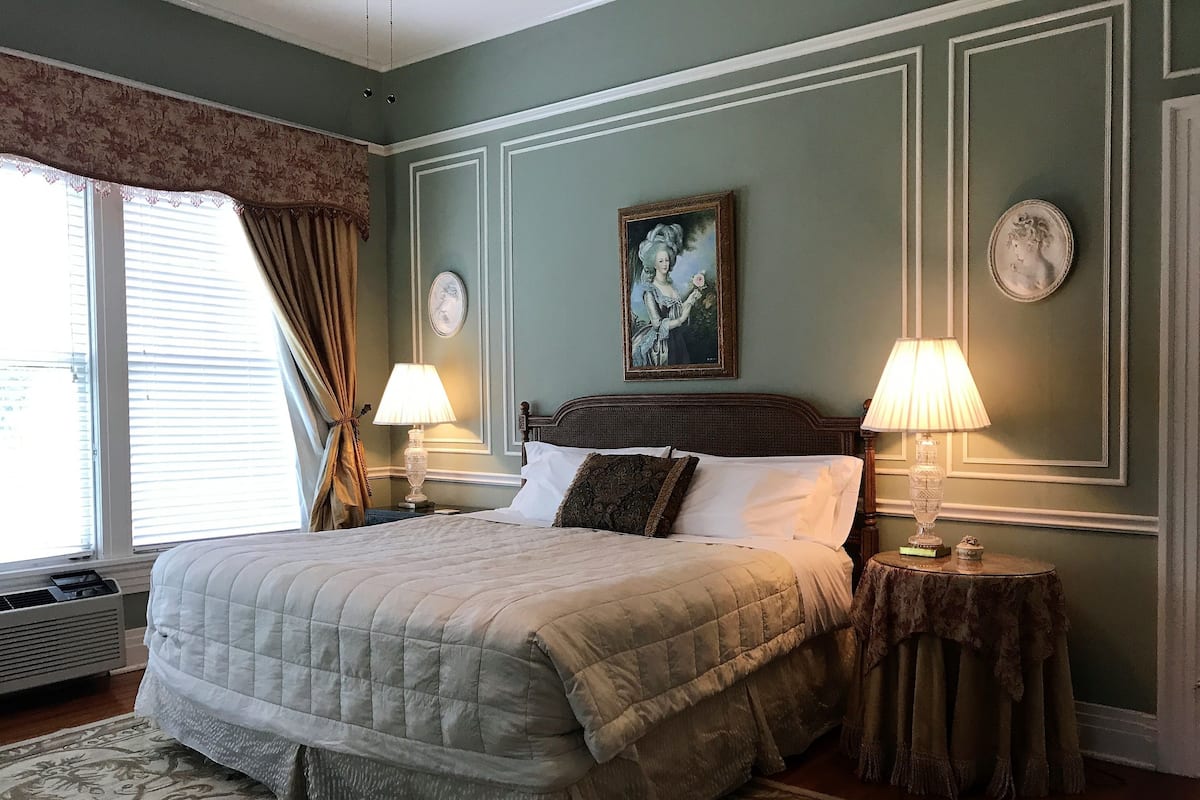 Un dormitorio ordenado con una cama grande, dos lámparas de noche, un reloj decorativo en la pared y una ventana con cortinas.
