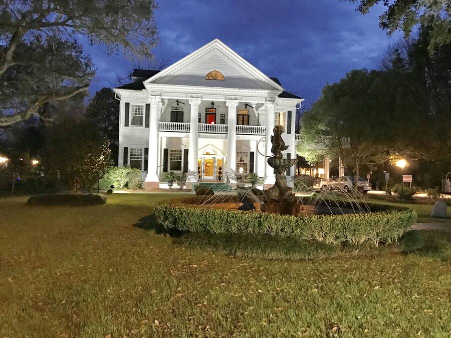A large, white, two-story building with a symmetrical facade, a central entrance with a portico, and a statue in the front garden.