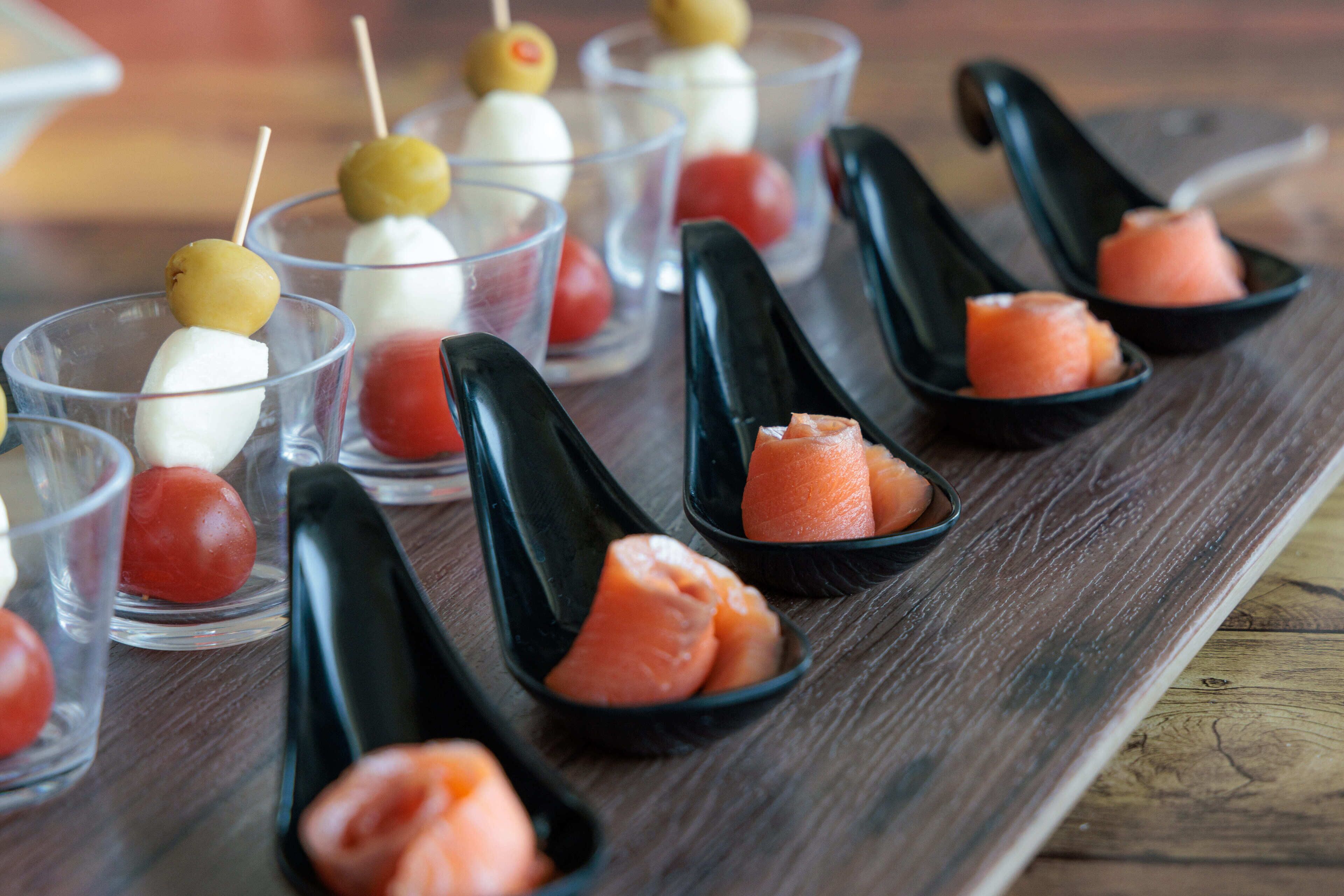 A wooden table with various food items, including skewers with olives and white cubes, and small black bowls with salmon.
