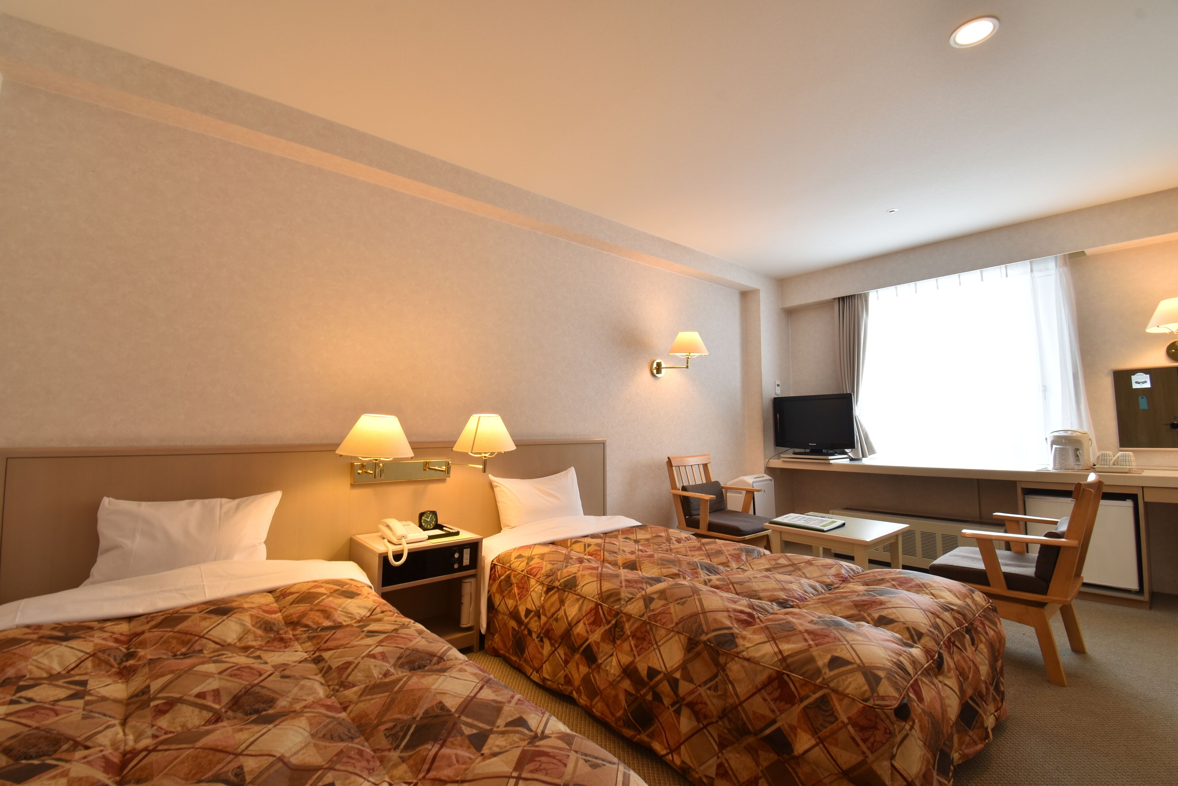 A hotel room with two beds, a desk, a chair, a television, and a window.