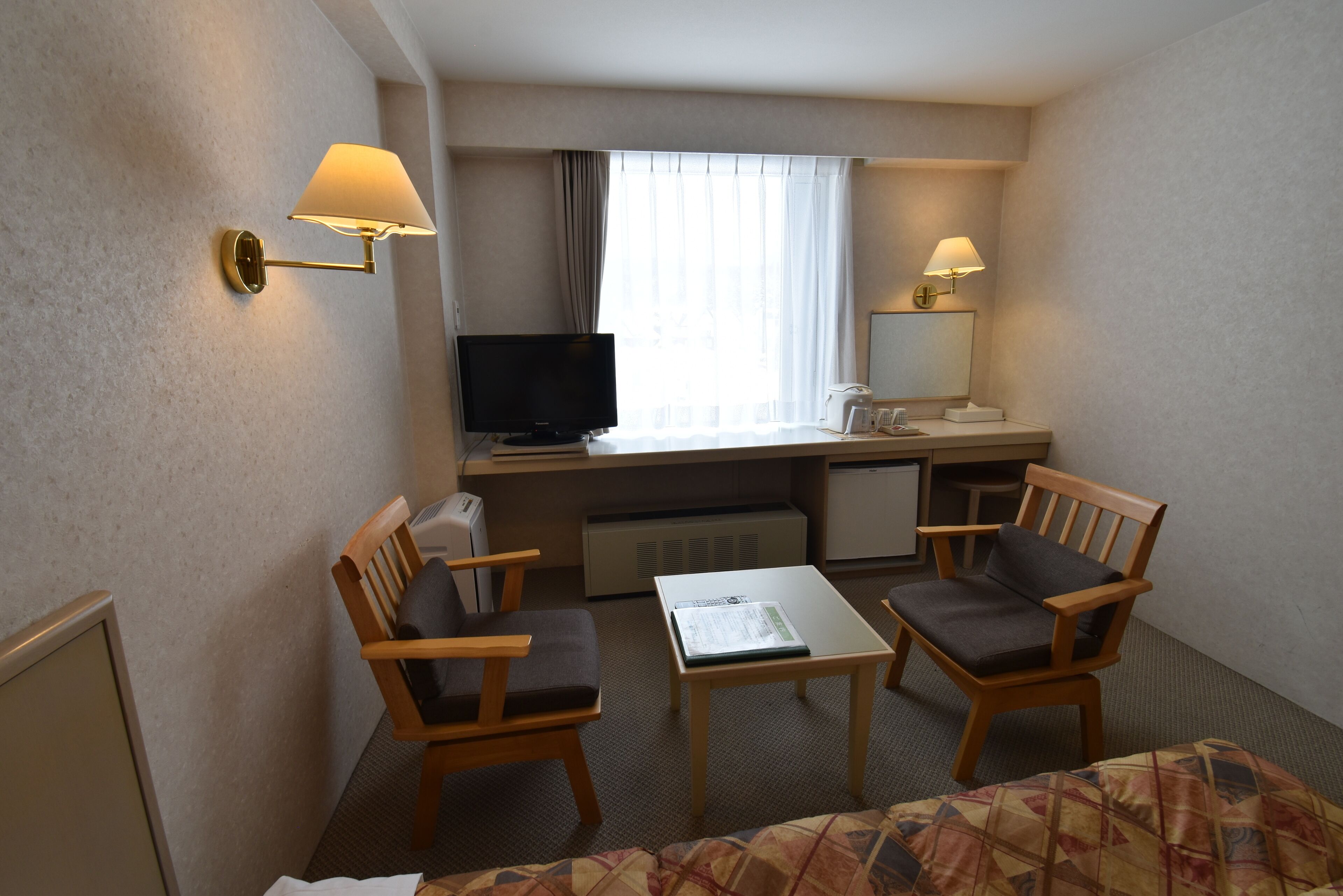 A hotel room with a desk, two chairs, a television, a bed, and a window with curtains.