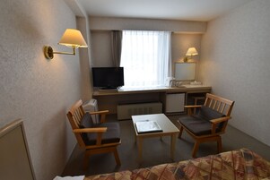 A hotel room with a desk, two chairs, a television, a bed, and a window with curtains.