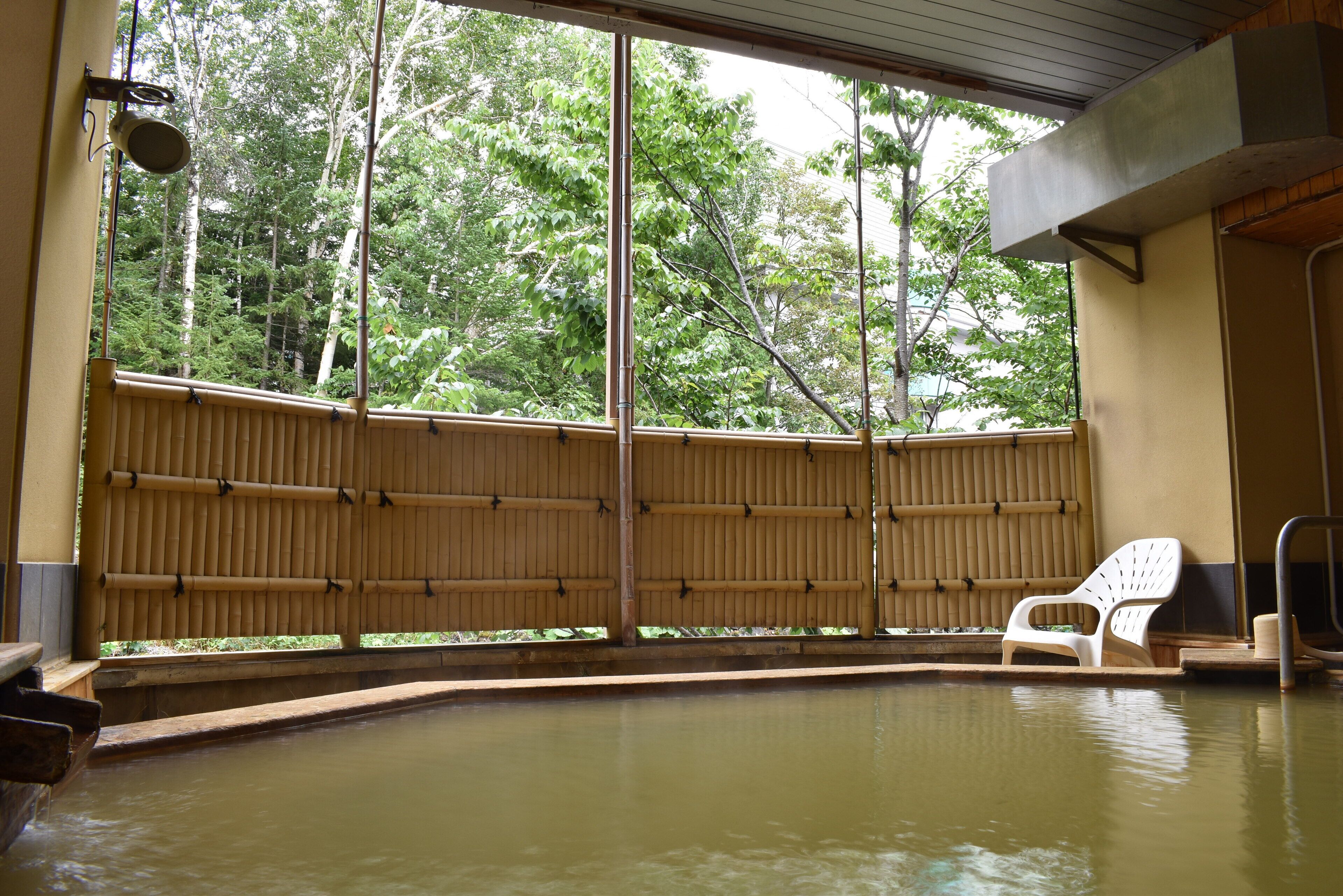 A traditional Japanese onsen with a wooden wall, a white plastic chair, and a view of a forested area.