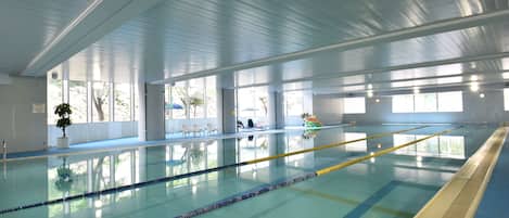 An indoor swimming pool with clear lanes, a blue floor, and large windows allowing natural light.
