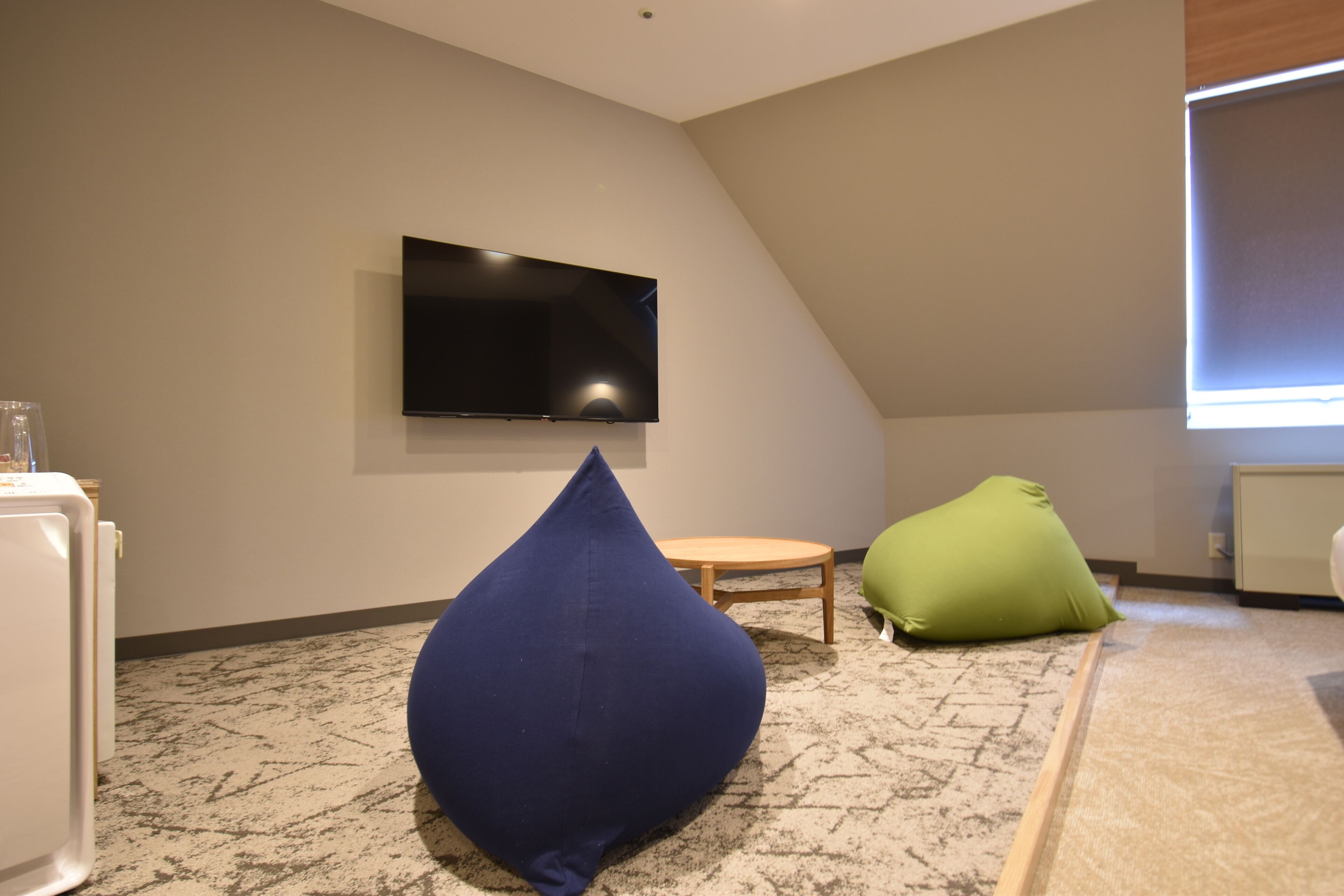 A room with a flat-screen TV mounted on the wall, a blue bean bag chair, a green bean bag chair, and a small wooden table.