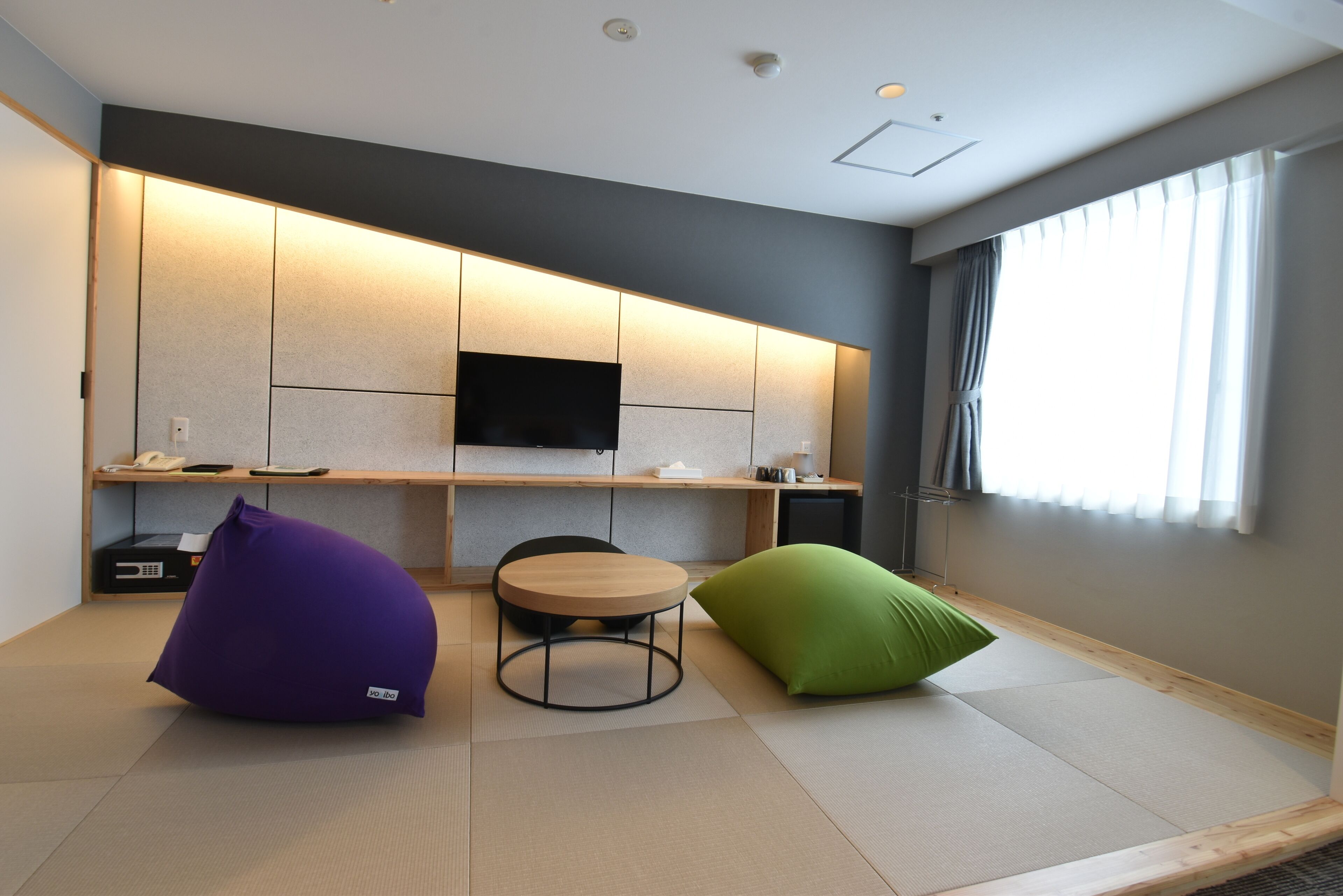 Habitació moderna amb sòl de tatami, una taula rodona, dos seients de bossa, un televisor de pantalla plana muntat a la paret i una finestra amb cortines.