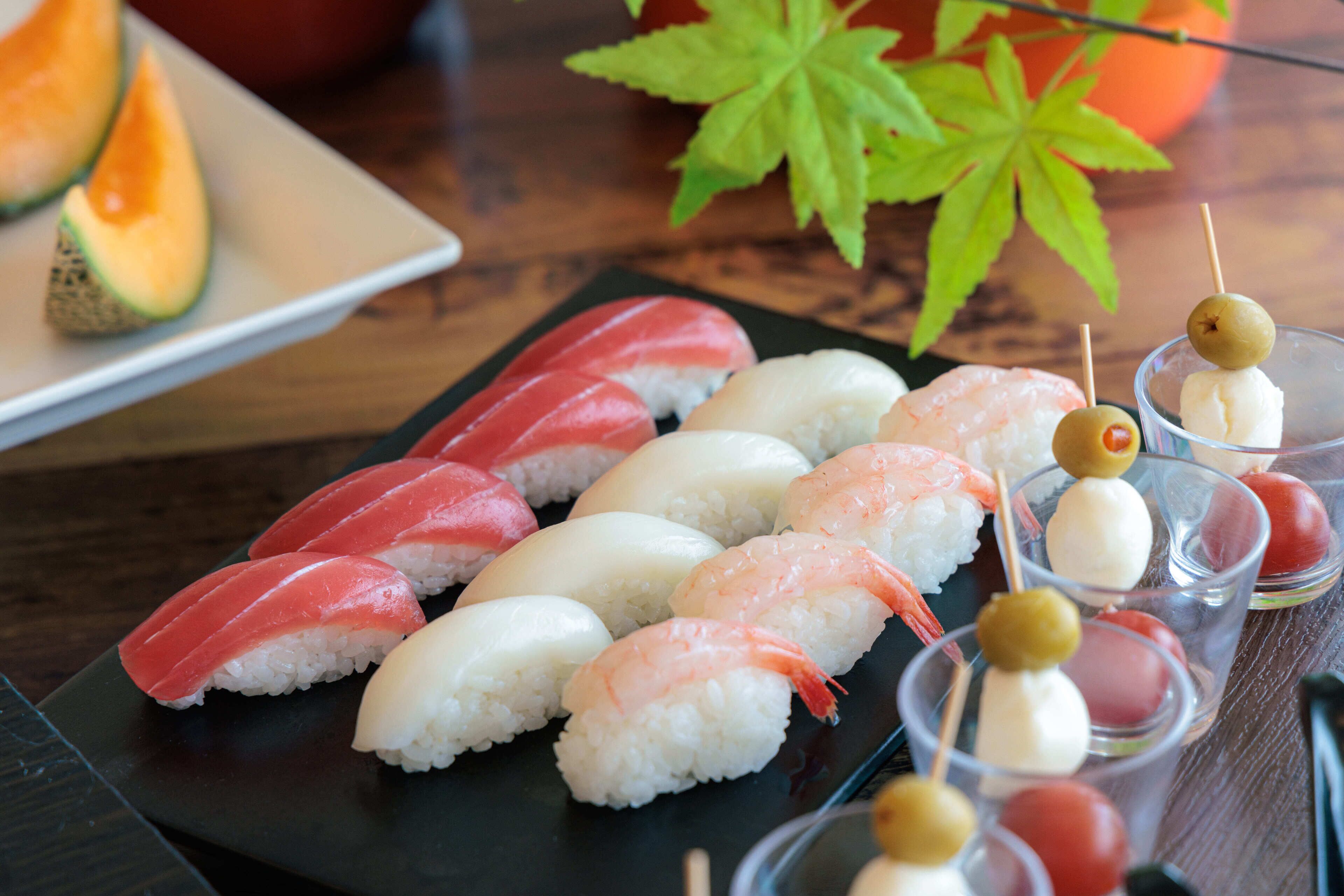 Verschiedene Sushi-Sorten auf einer schwarzen Platte, darunter Nigiri und Sashimi, dazu ein kleines Glas Sake und Kirschtomaten am Spieß.