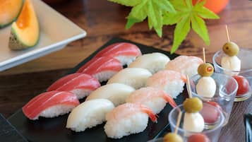Verschiedene Sushi-Sorten auf einer schwarzen Platte, darunter Nigiri und Sashimi, dazu ein kleines Glas Sake und Kirschtomaten am Spieß.