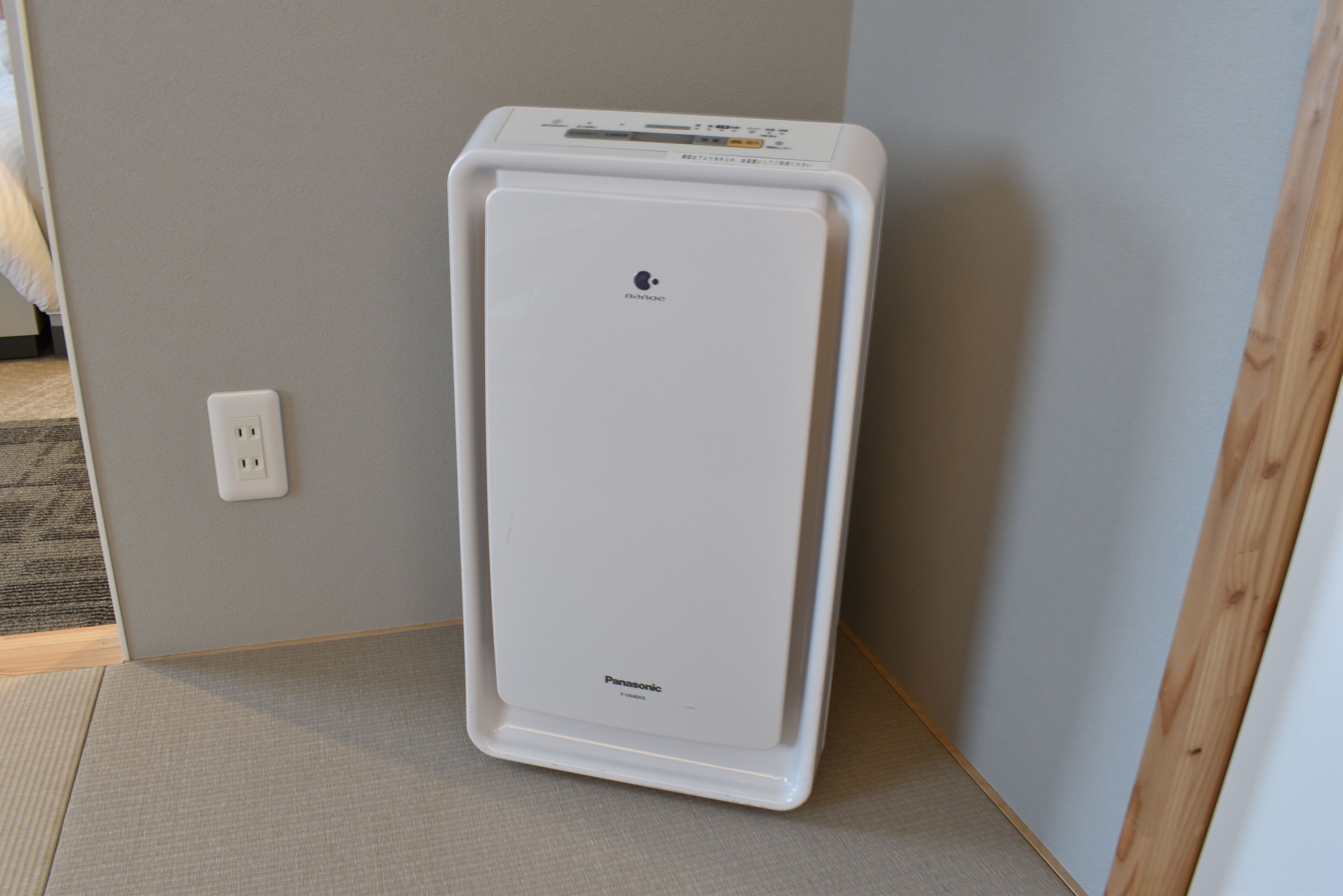 A white portable heater in a corner of a room with a light gray wall and a wooden floor.