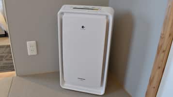 A white portable heater in a corner of a room with a light gray wall and a wooden floor.