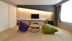 Una habitación moderna con suelo de tatami, una mesa redonda de madera, dos pufs, un televisor de pantalla plana y un estante empotrado con una cafetera.