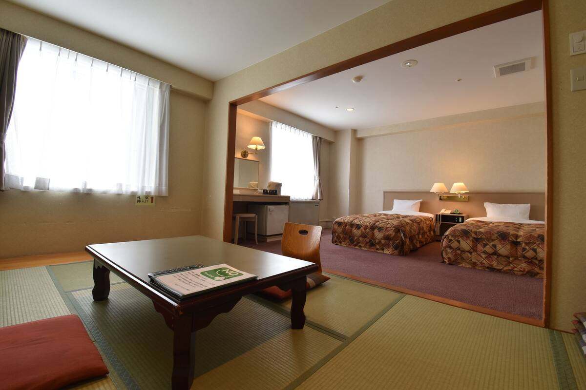Habitación de hotel con tatamis, una mesa de madera, dos camas con ropa de cama estampada, un escritorio con una silla y una pequeña kitchenette.