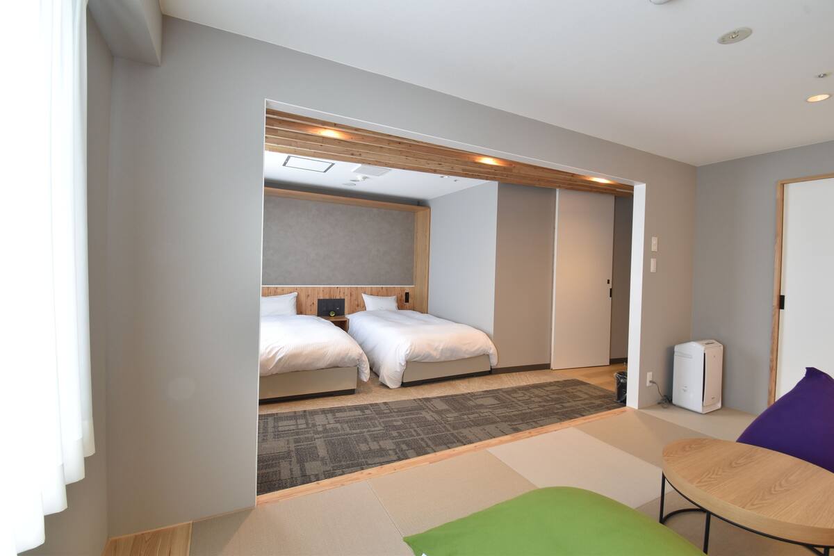 Habitación de hotel con dos camas, una mesita, una silla y un cubo de basura visible.