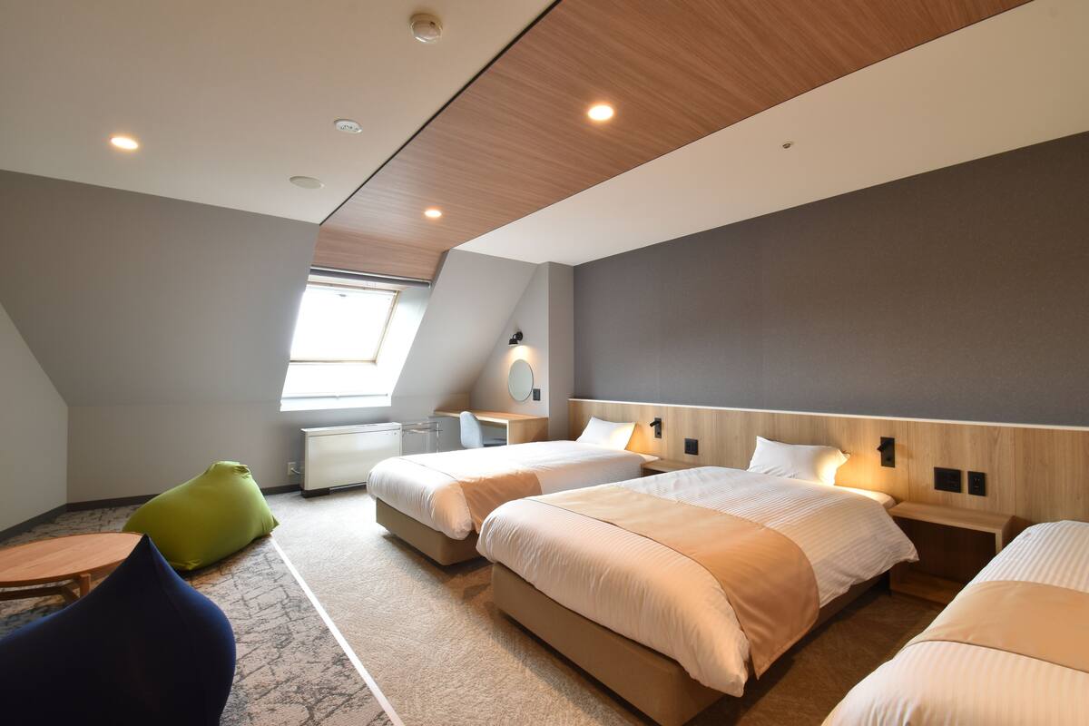 Una habitación de hotel moderna con tres camas, un escritorio, una silla y un ventanal.