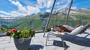 A balcony with a view of a mountain range, a chair, a table with a glass of beer, and a potted plant with flowers. - Alpenhotel Laurin (Soelden)