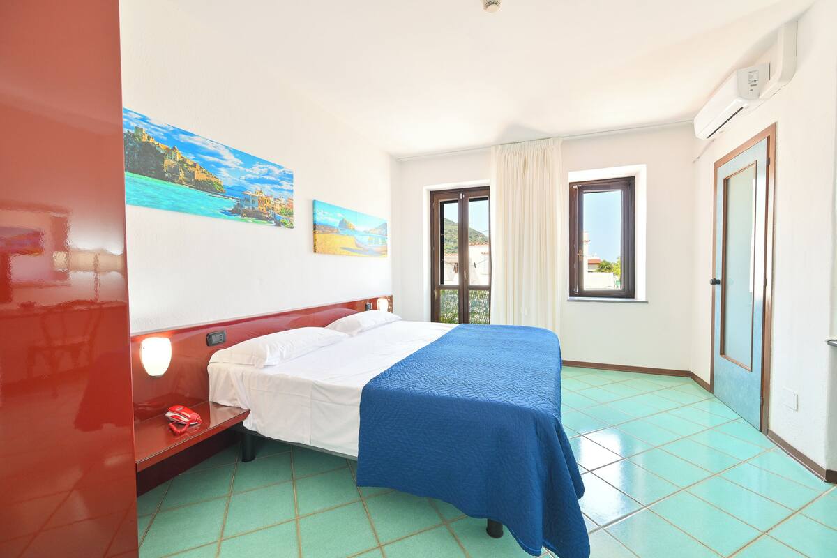 A hotel room with a bed, a window with curtains, a door, and a wall with two paintings.