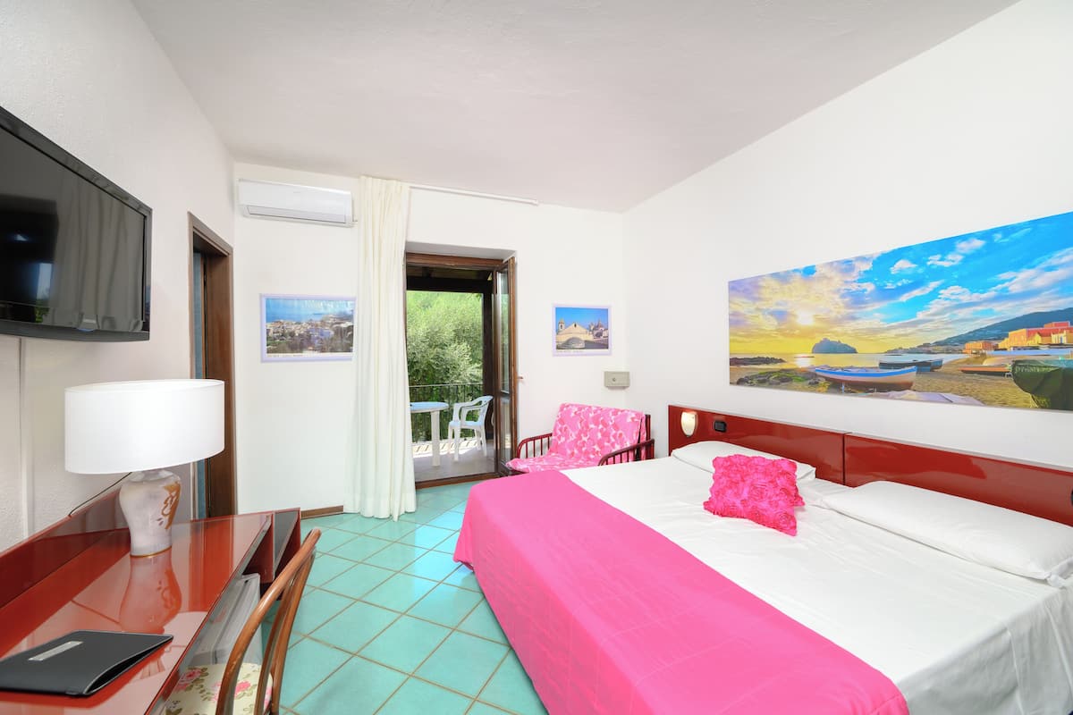 A hotel room with a bed, a desk with a lamp, a TV, and a balcony with chairs.