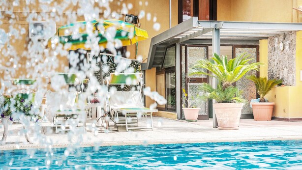 Un área de piscina con sillas de descanso y sombrillas, una zona de estar cubierta y plantas en macetas.