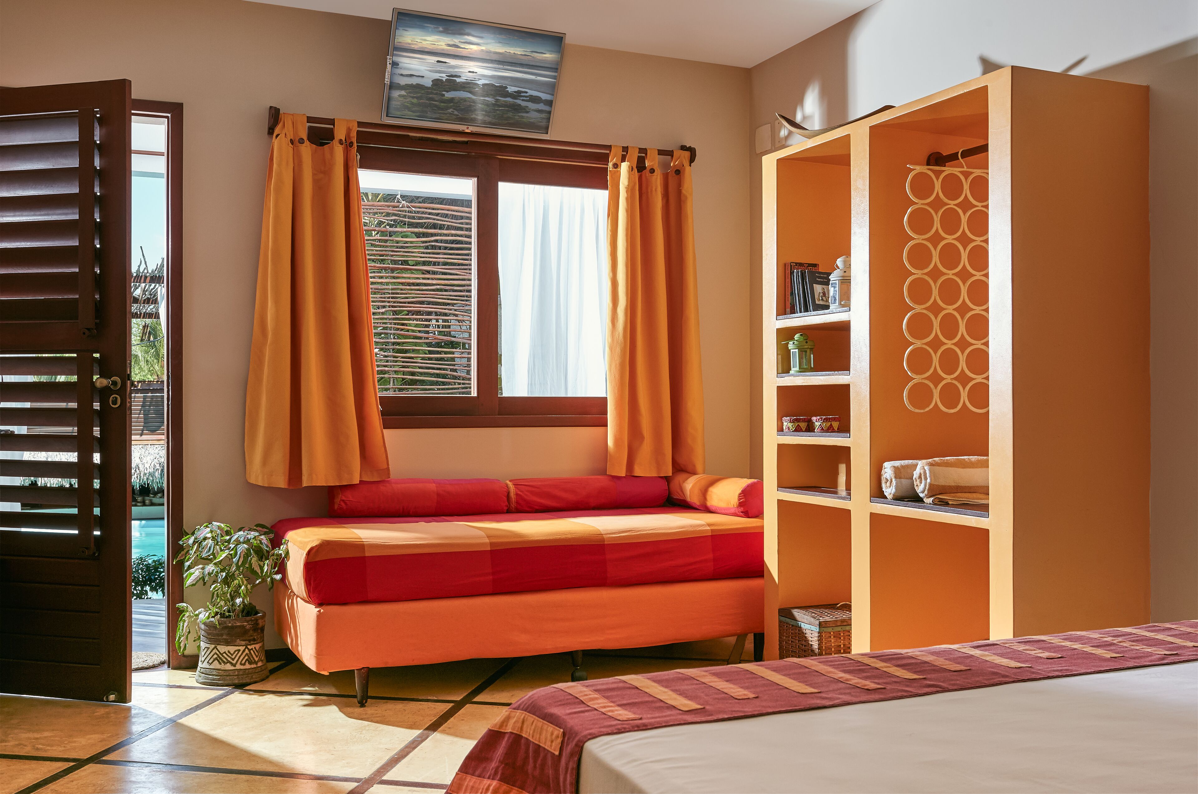 Quarto com cama, guarda-roupa, janela com cortinas, planta em vaso e prateleira com objetos.