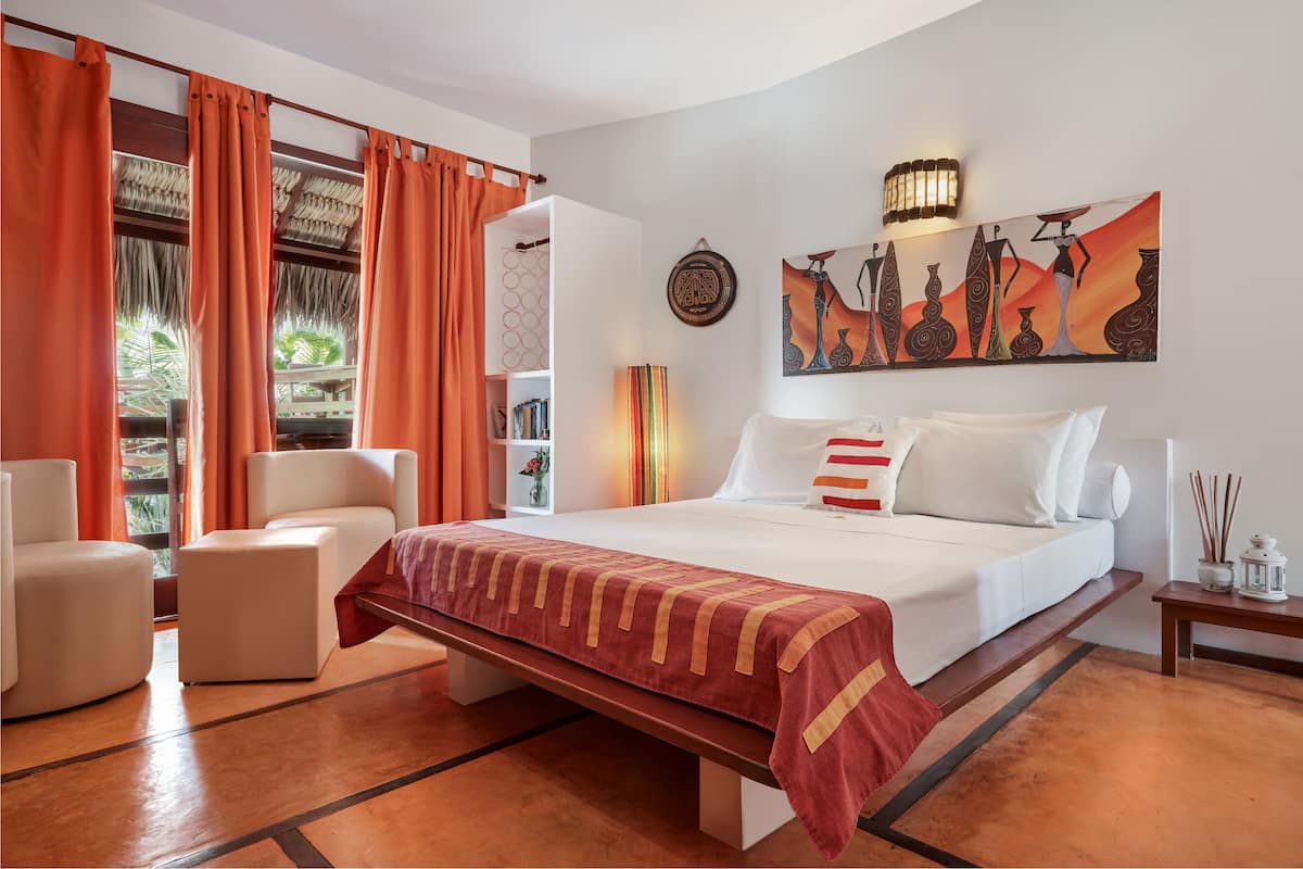 Quarto de hotel com cama, duas cadeiras, mesa de cabeceira, estante de livros e uma janela grande com cortinas.