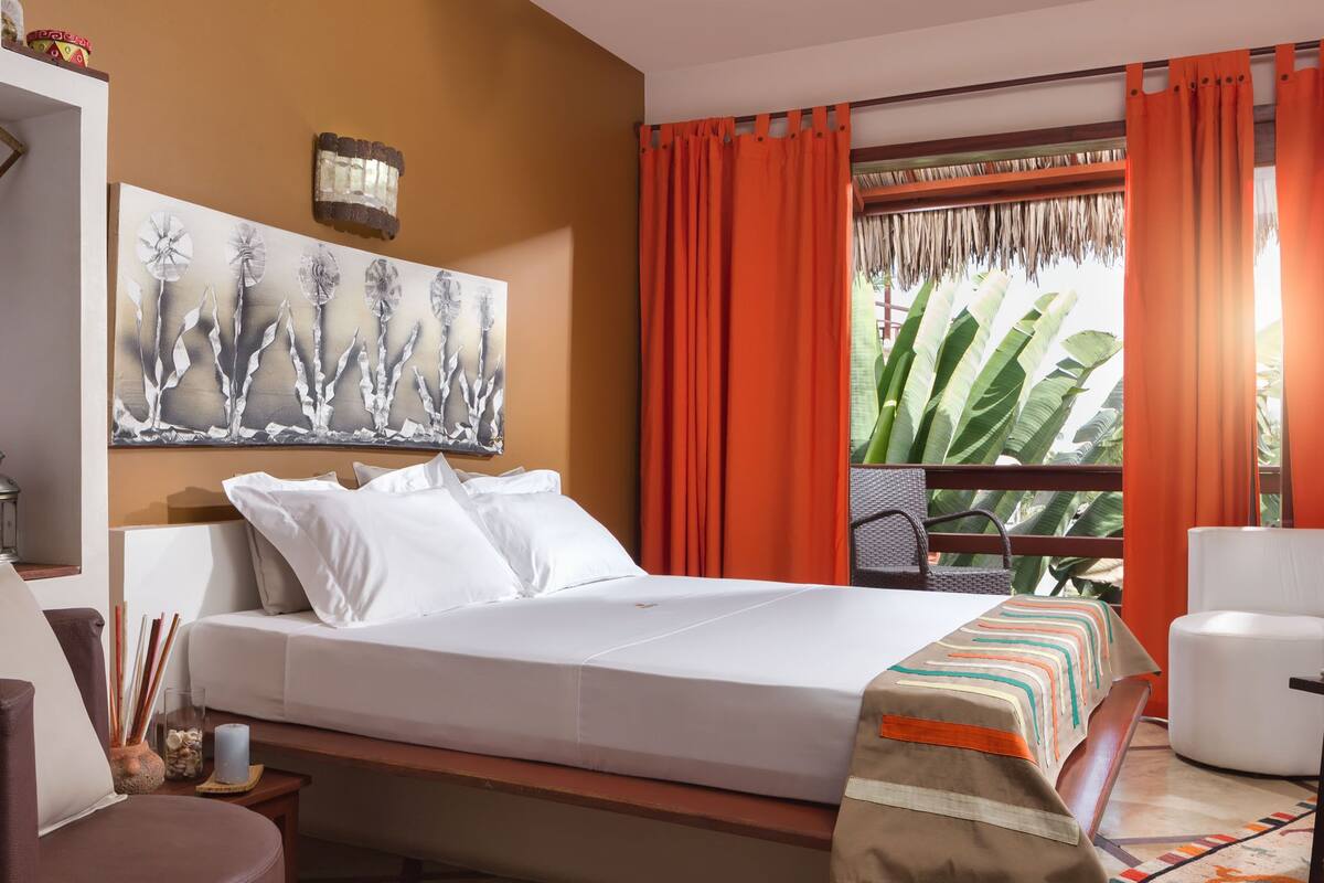 Quarto de hotel com cama, poltrona, escrivaninha, janela grande com cortinas e um quadro na parede.