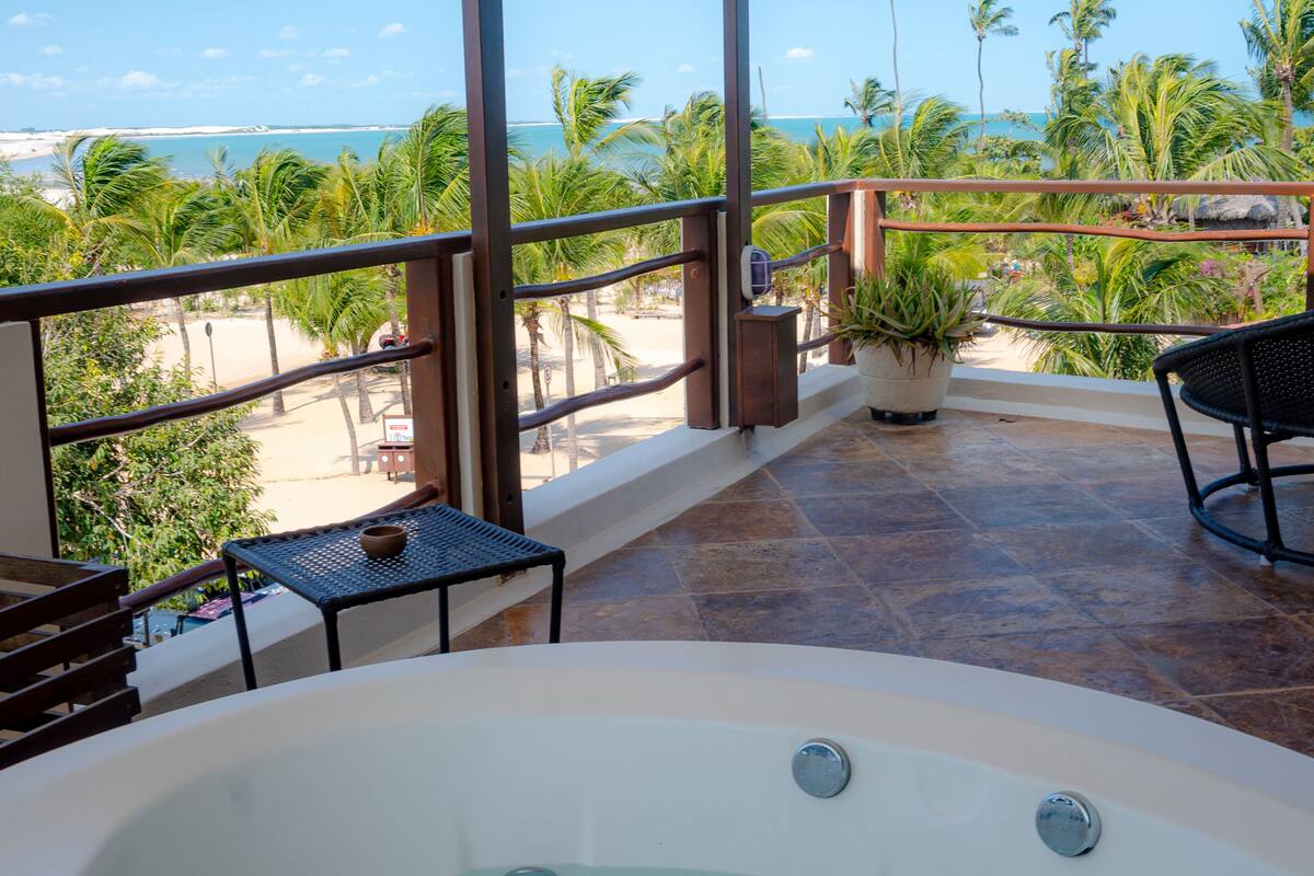 Uma varanda com vista para a praia e oceano, uma cadeira de vime preto, uma mesa pequena com uma tigela e uma planta em vaso.