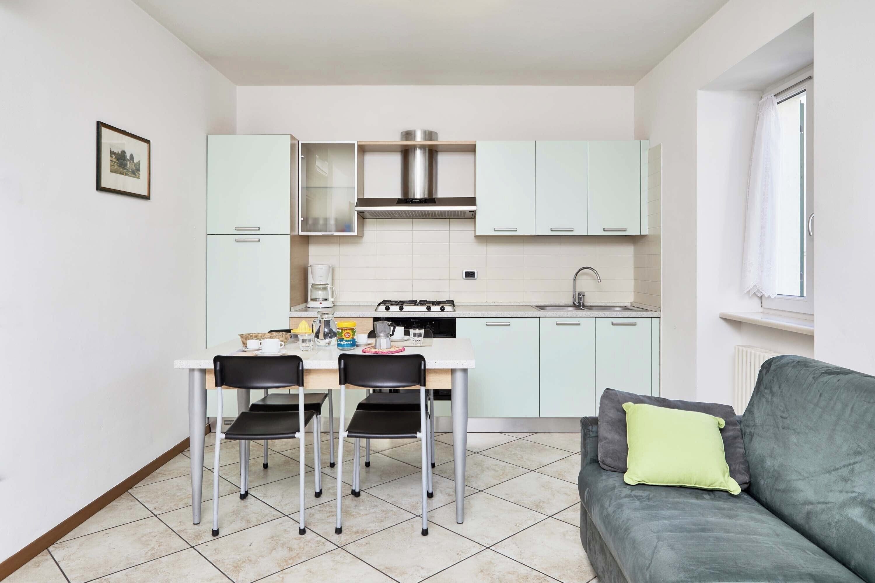A compact kitchen with white cabinets, a dining table with chairs, and a grey sofa with a green pillow.
