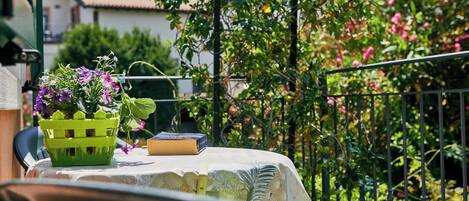 Ein Balkon mit Tisch, bedeckt mit einer Spitzentischdecke, einem grünen Korb mit Blumen und einem Buch.