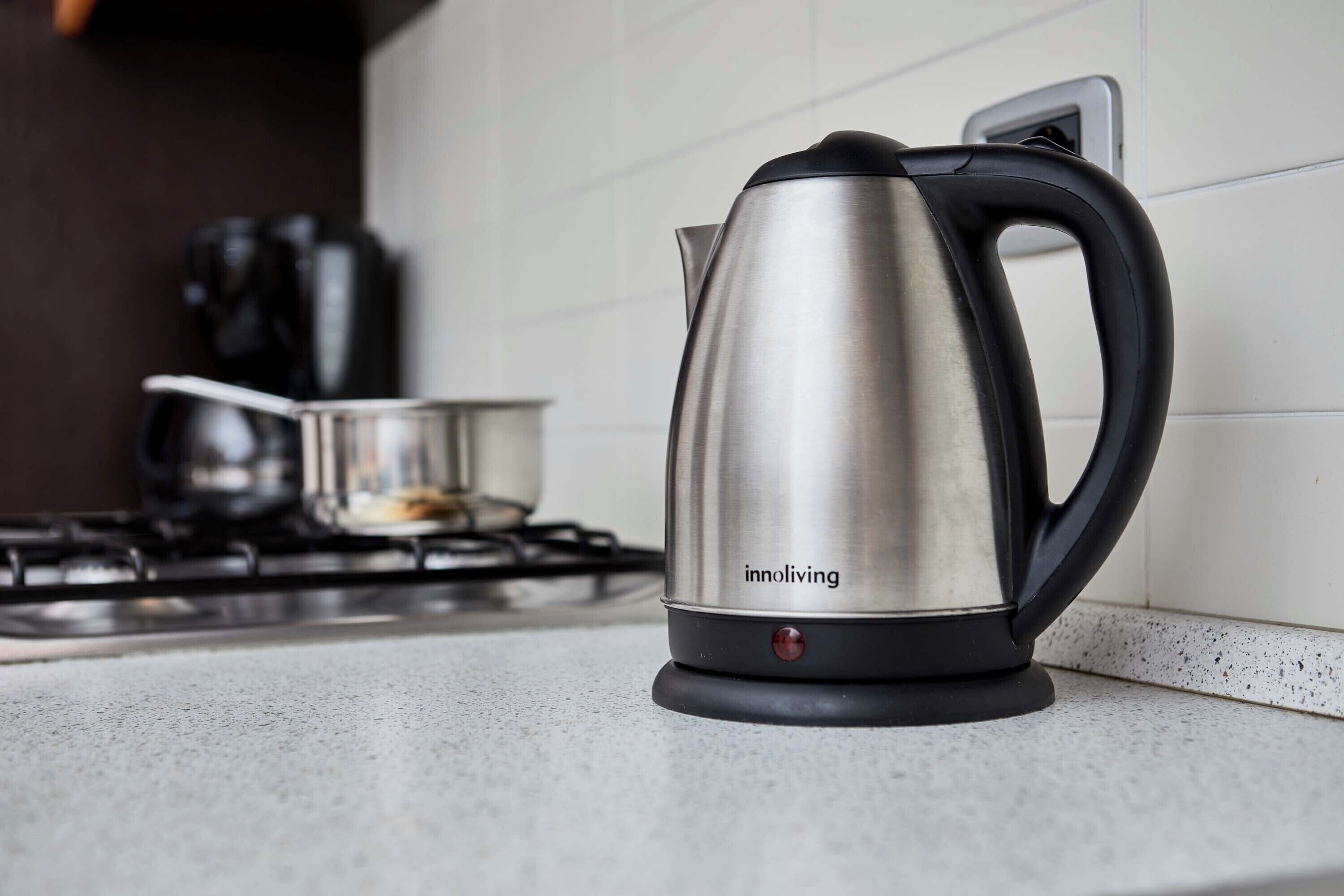 Una tetera de acero inoxidable con asa negra y un botón rojo, sobre una encimera de cocina.