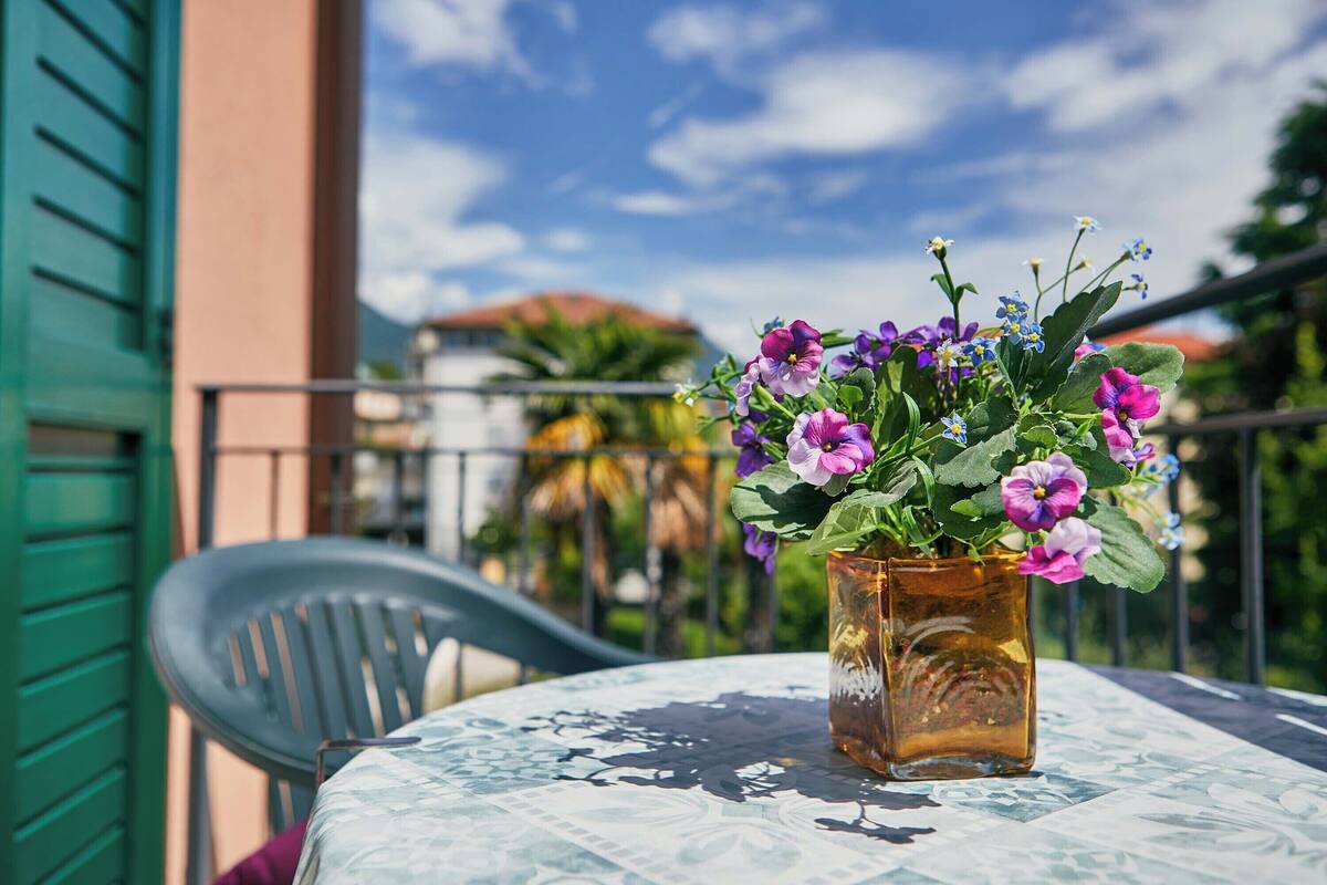 A table with a vase of flowers on a balcony with a green chair and a railing.
